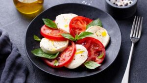 Ensalada Caprese con queso fresco: Rodajas de tomate y queso fresco, alternadas y decoradas con hojas de albahaca, aderezadas con aceite de oliva y pimienta negra.