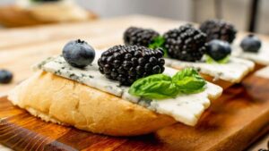 Una rebanada de pan crujiente con una generosa porción de queso azul, adornada con moras frescas y una hoja de albahaca, ofrece una combinación de sabores y texturas que deleitan el paladar.