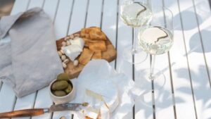 Un plato de queso camembert, pistachos y galletas saladas, acompañado de aceitunas verdes y dos copas de vino blanco, listo para una experiencia gastronómica.